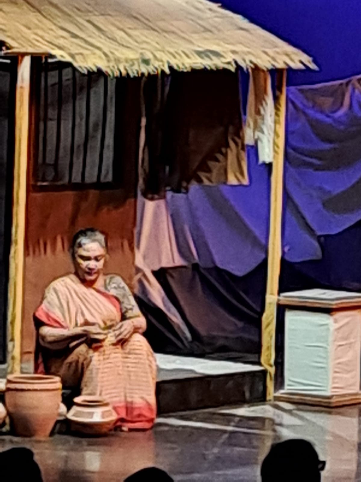 Performer seated beside clay pots outside a hut set